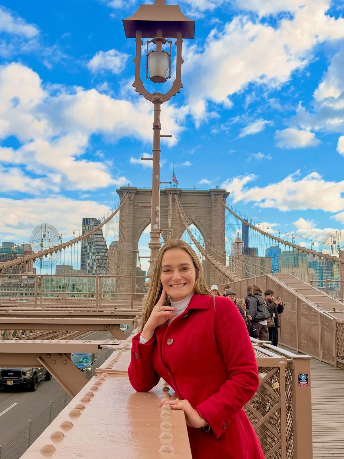 Lore de casaco vermelho apoiada no parapeito da Ponte do Brooklyn, com os arranha-céus de Manhattan ao fundo