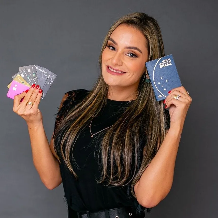 Lore em foto de estúdio, sorrindo com um leque de cartões de crédito numa das mãos e o passaporte brasileiro na outra